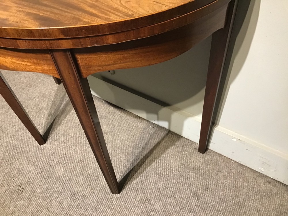 A semi-circular antique mahogany demi-lune card table featuring four square tapered legs and a patterned flame mahogany veneered top surface. View IV