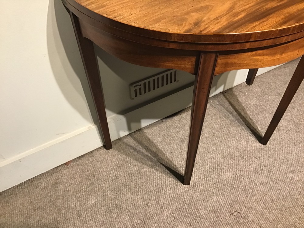 A semi-circular antique mahogany demi-lune card table featuring four square tapered legs and a patterned flame mahogany veneered top surface. View V