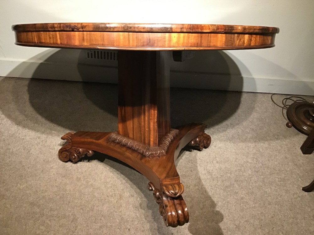 A circular William IV Goncalo Alves tilt-top centre table tilted vertically to show the figured timber grain above a triform base with carved scroll feet. View IV