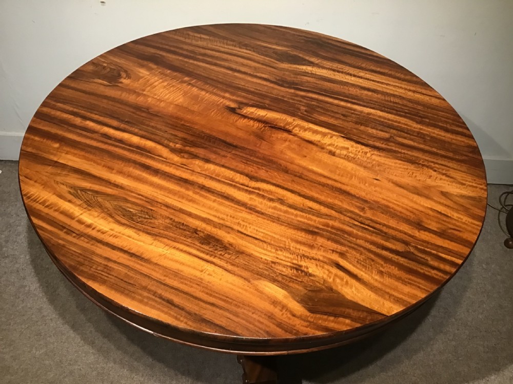 A circular William IV Goncalo Alves tilt-top centre table tilted vertically to show the figured timber grain above a triform base with carved scroll feet. View IX