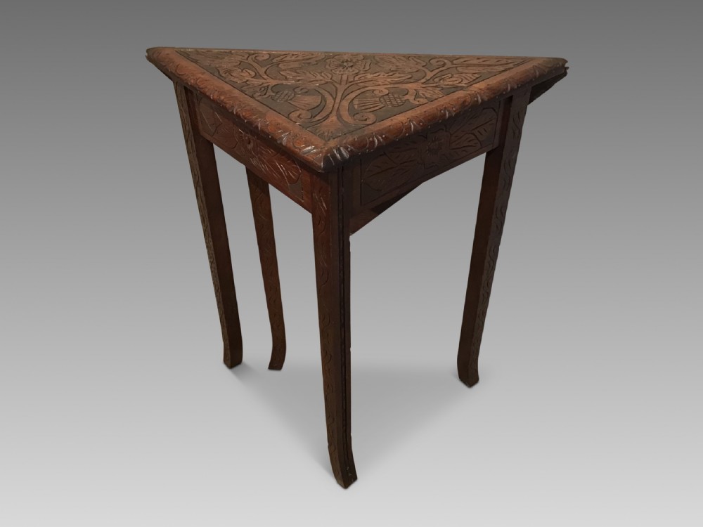 A triangular dark carved wooden corner table featuring intricate relief carvings of leaves and flowers on its top surface and legs.