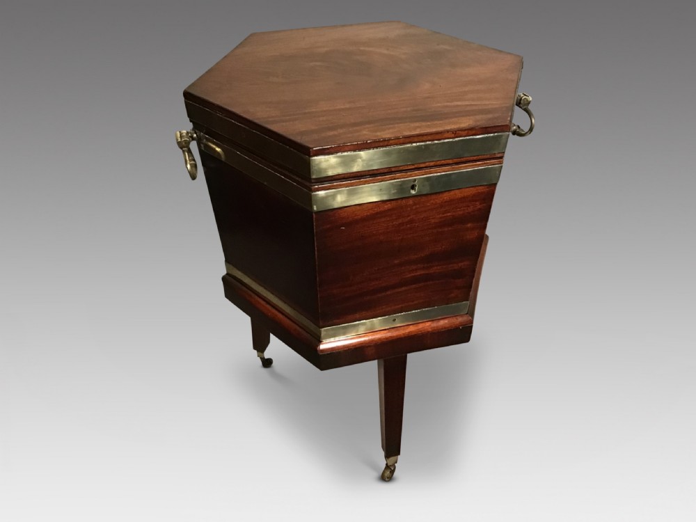 A hexagonal dark mahogany Georgian wine cooler featuring brass binding straps, side handles, and tapered legs on brass castors.
