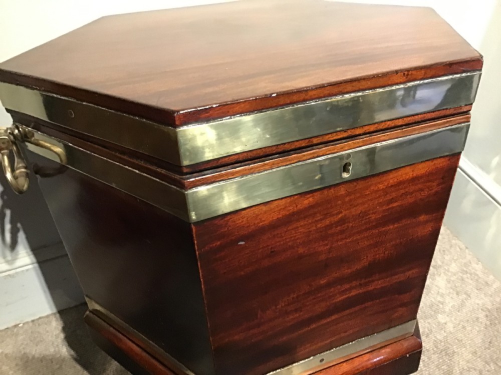 A hexagonal dark mahogany Georgian wine cooler featuring brass binding straps, side handles, and tapered legs on brass castors. View II