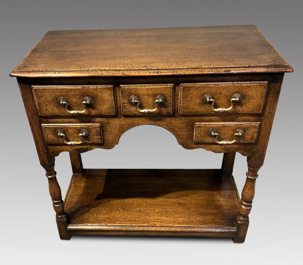 An antique warm brown oak side table featuring five drawers with brass handles, a central arched apron, turned front legs, and a lower potboard shelf.
