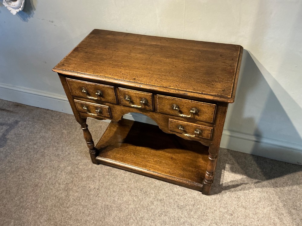 An antique warm brown oak side table featuring five drawers with brass handles, a central arched apron, turned front legs, and a lower potboard shelf. View II