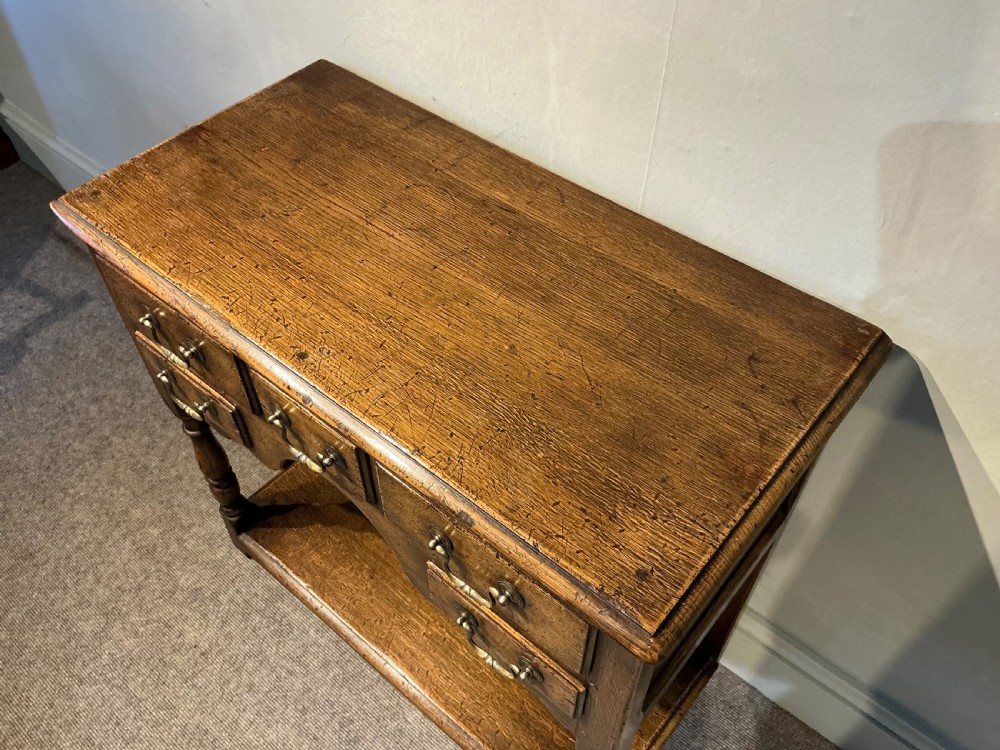 An antique warm brown oak side table featuring five drawers with brass handles, a central arched apron, turned front legs, and a lower potboard shelf. View III