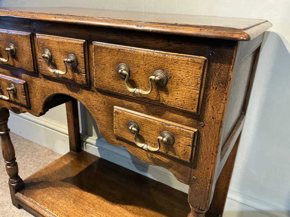 An antique warm brown oak side table featuring five drawers with brass handles, a central arched apron, turned front legs, and a lower potboard shelf. View IV