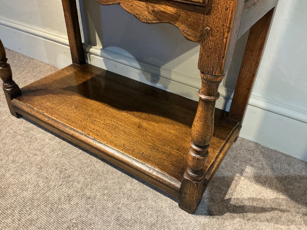 An antique warm brown oak side table featuring five drawers with brass handles, a central arched apron, turned front legs, and a lower potboard shelf. View V