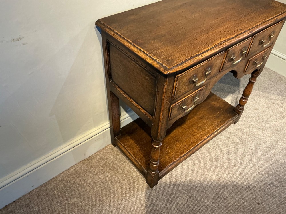 An antique warm brown oak side table featuring five drawers with brass handles, a central arched apron, turned front legs, and a lower potboard shelf. View VI