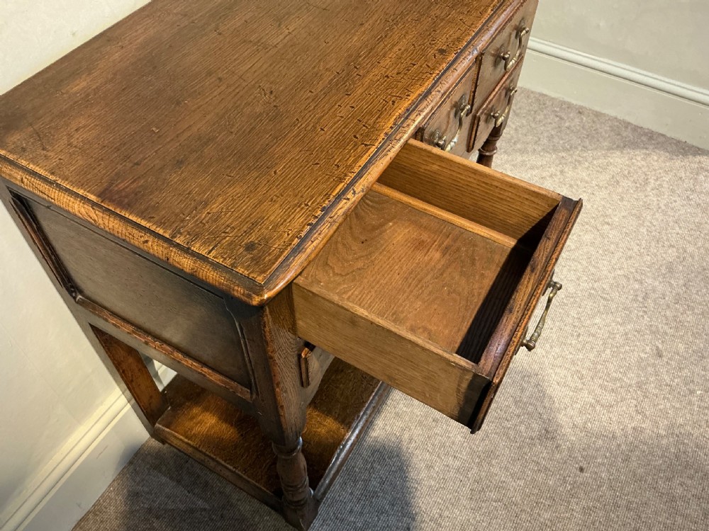 An antique warm brown oak side table featuring five drawers with brass handles, a central arched apron, turned front legs, and a lower potboard shelf. View VII