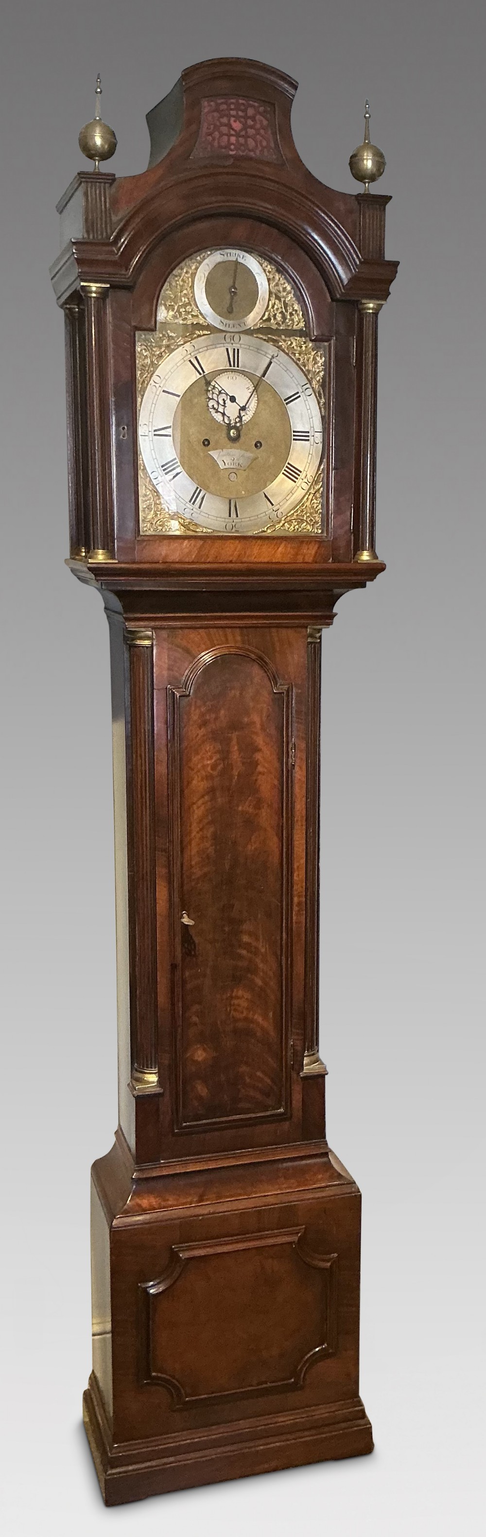 An antique Georgian longcase clock crafted from dark flame mahogany, featuring an arched hood with brass finials and an engraved brass dial.