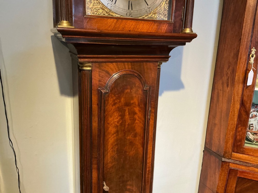 An antique Georgian longcase clock crafted from dark flame mahogany, featuring an arched hood with brass finials and an engraved brass dial. View IV