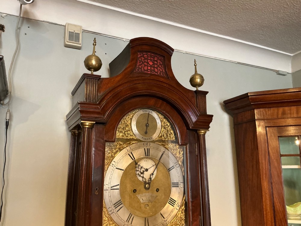 An antique Georgian longcase clock crafted from dark flame mahogany, featuring an arched hood with brass finials and an engraved brass dial. View VI