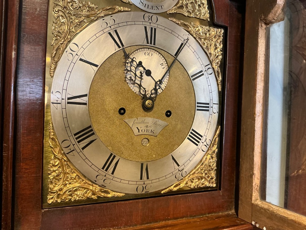 An antique Georgian longcase clock crafted from dark flame mahogany, featuring an arched hood with brass finials and an engraved brass dial. View VII
