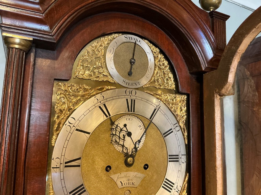 An antique Georgian longcase clock crafted from dark flame mahogany, featuring an arched hood with brass finials and an engraved brass dial. View VIII