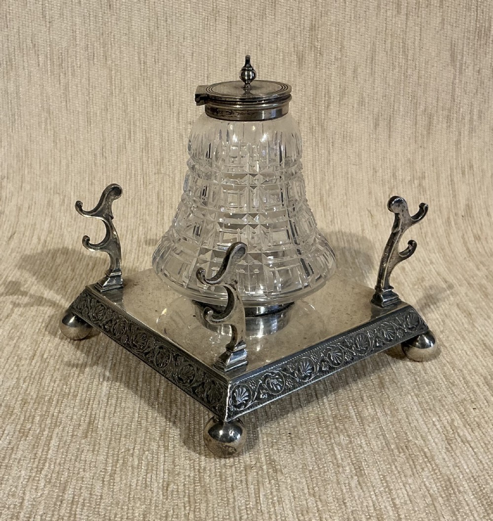 A square silver-plated inkstand featuring embossed borders, ball feet, integrated pen rests, and a central cut-glass inkwell with a hinged silver cap.