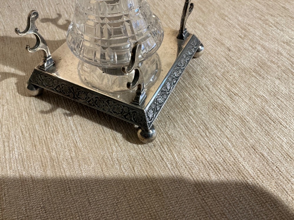 A square silver-plated inkstand featuring embossed borders, ball feet, integrated pen rests, and a central cut-glass inkwell with a hinged silver cap. View II