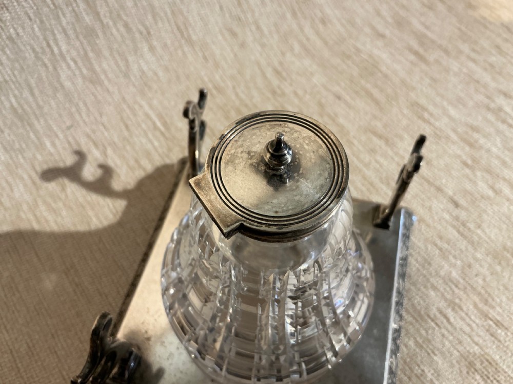 A square silver-plated inkstand featuring embossed borders, ball feet, integrated pen rests, and a central cut-glass inkwell with a hinged silver cap. View III