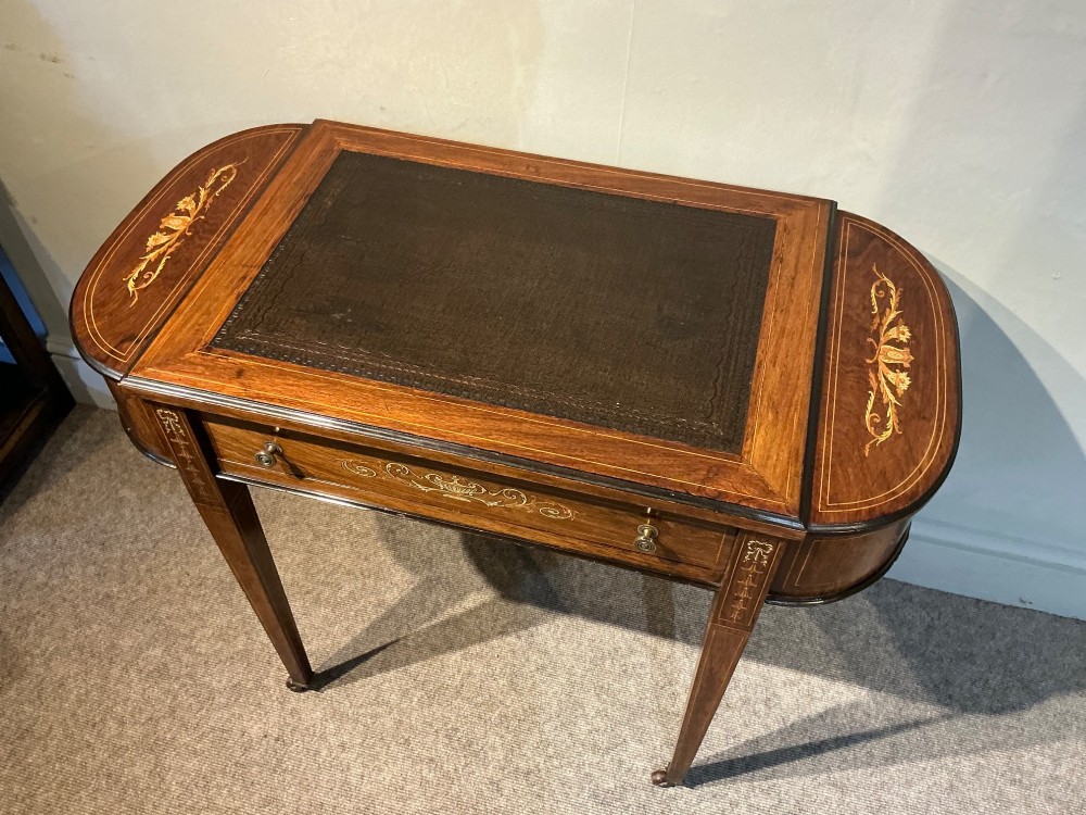 An antique inlaid rosewood writing table featuring a dark leather inset surface, classical urn marquetry, a single drawer, and square tapered legs on brass castors. View III