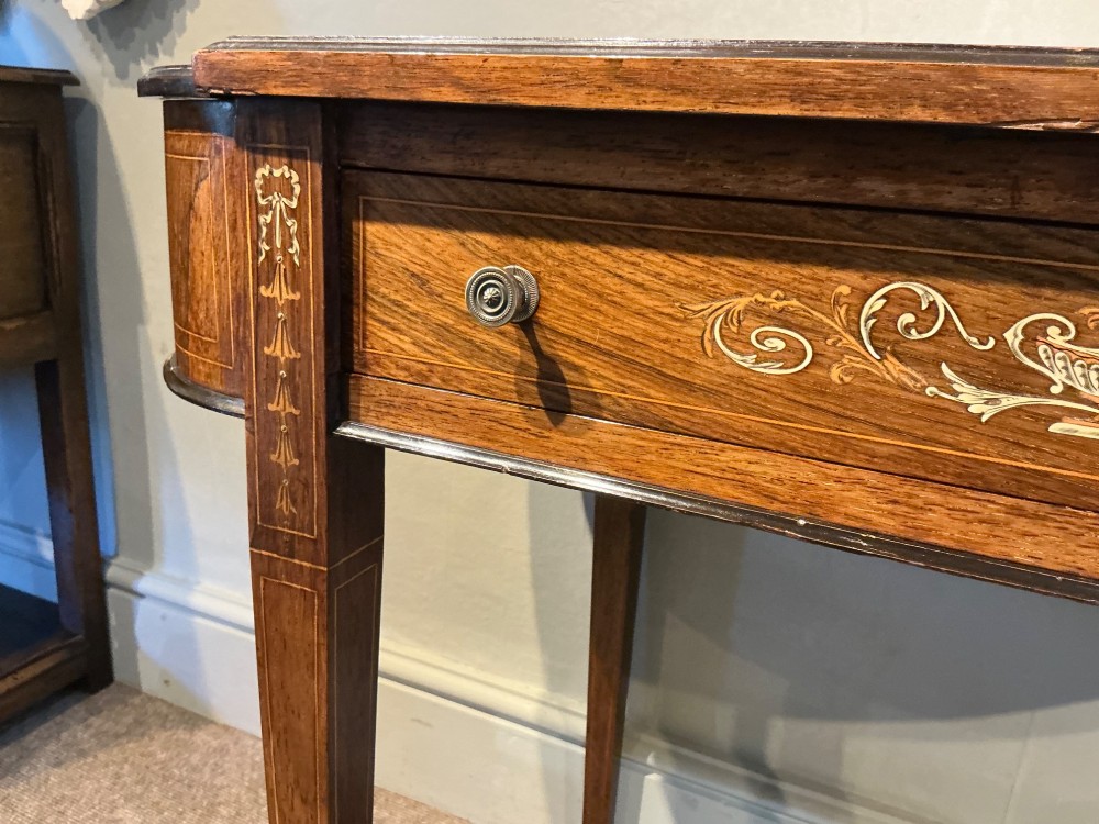 An antique inlaid rosewood writing table featuring a dark leather inset surface, classical urn marquetry, a single drawer, and square tapered legs on brass castors. View VII