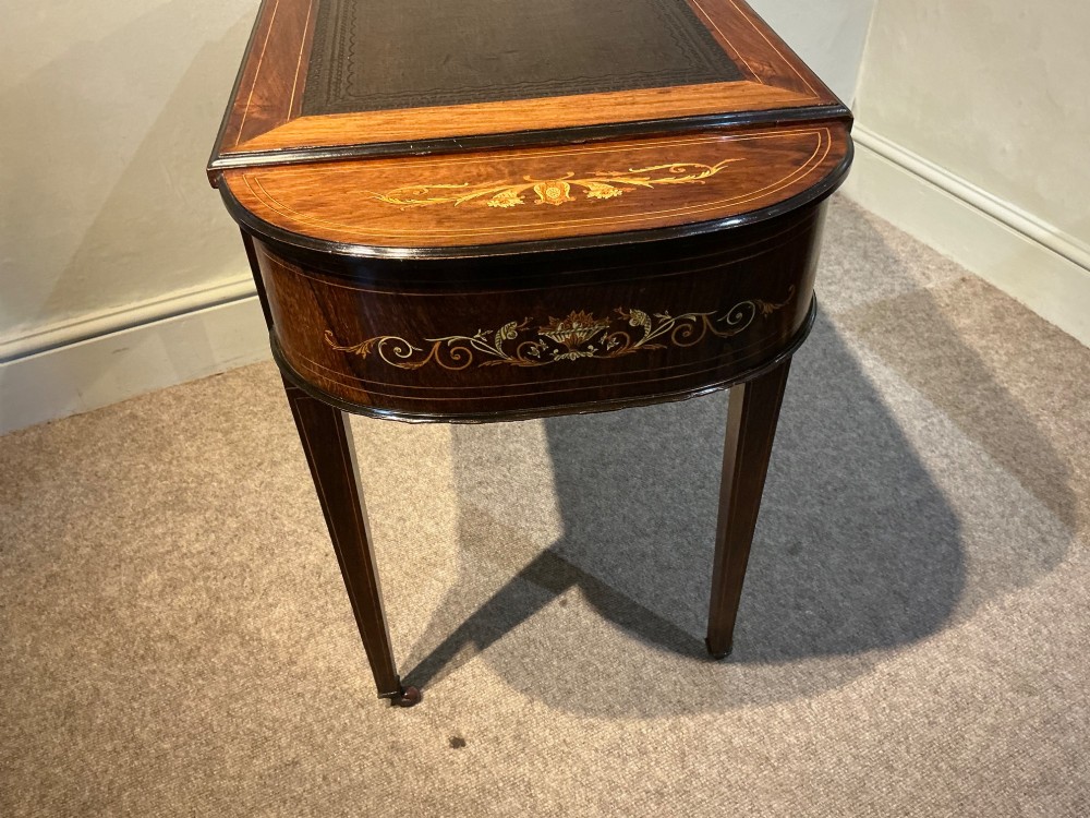 An antique inlaid rosewood writing table featuring a dark leather inset surface, classical urn marquetry, a single drawer, and square tapered legs on brass castors. View X