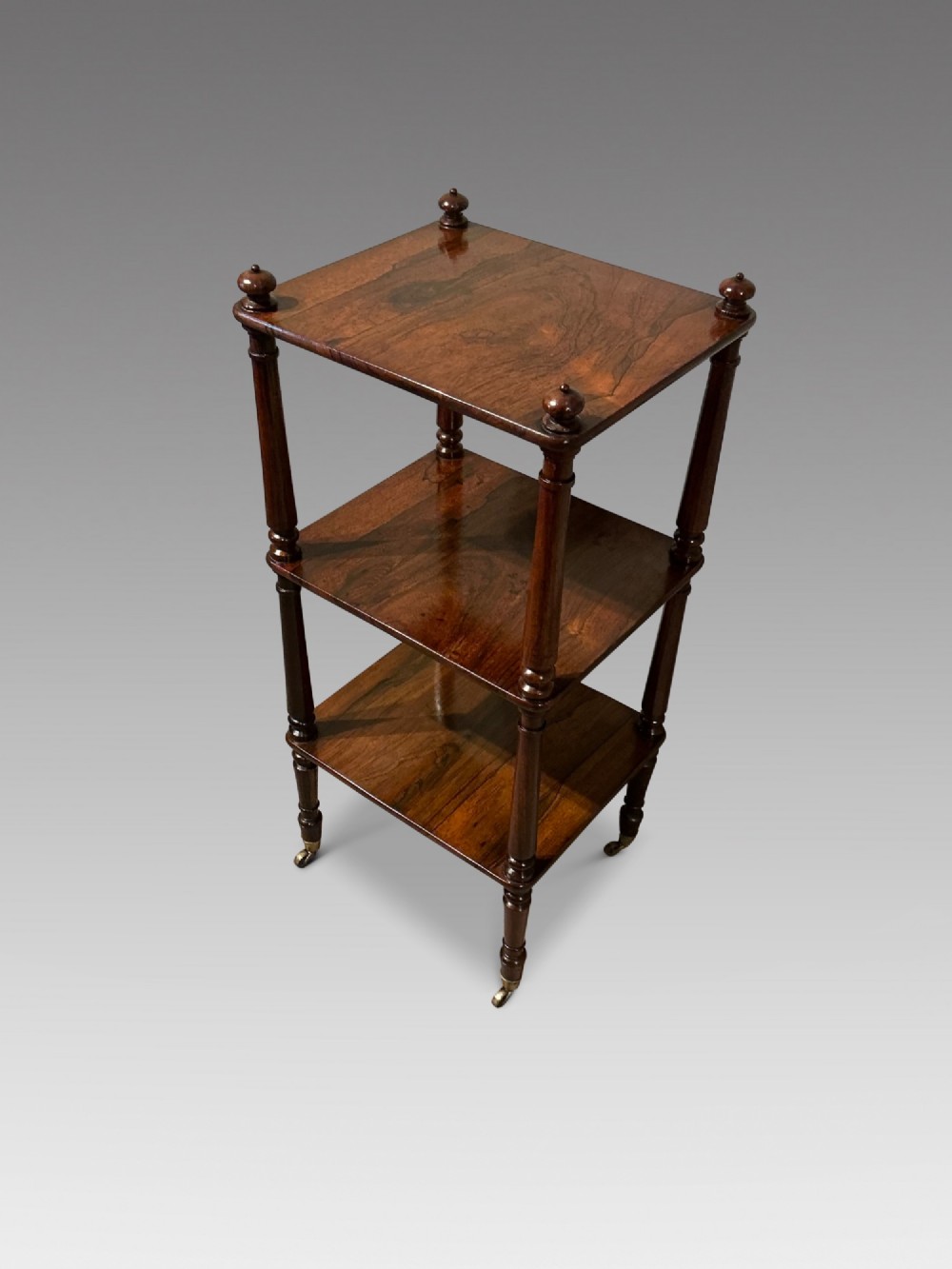A three-tiered dark rosewood Regency etagere featuring turned supports, corner finials, and brass castors.