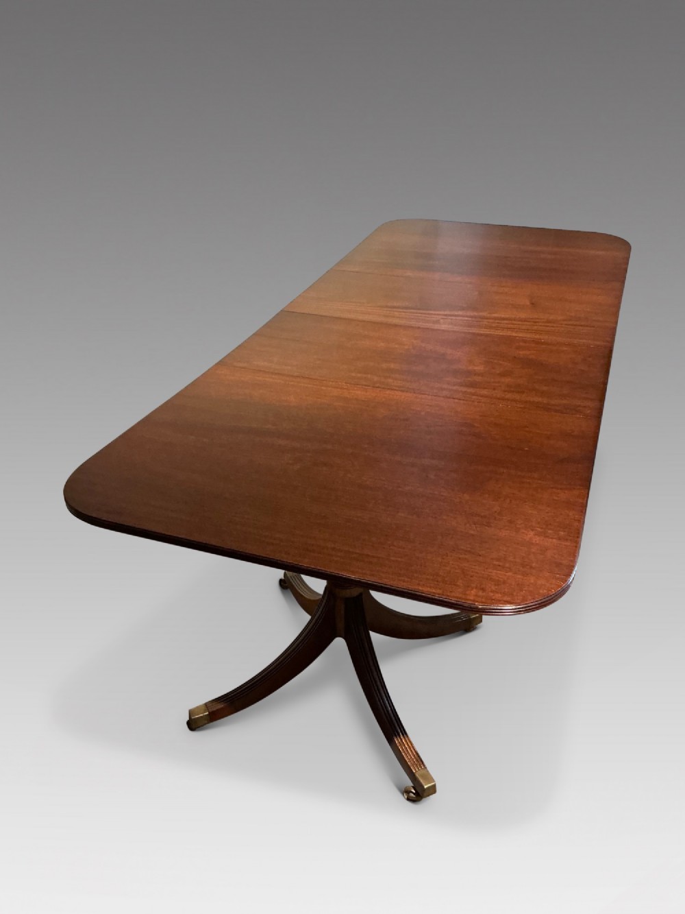 A mahogany twin pedestal dining table featuring rounded rectangular top corners, splayed tripod legs, and brass castors.