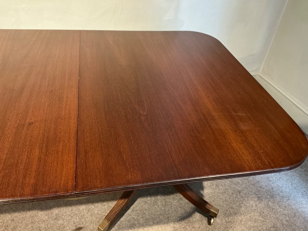 A mahogany twin pedestal dining table featuring rounded rectangular top corners, splayed tripod legs, and brass castors. View VI