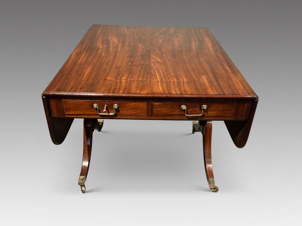 An antique Georgian mahogany sofa table with two drop leaves, two front drawers with brass handles, and splayed legs resting on brass castors.