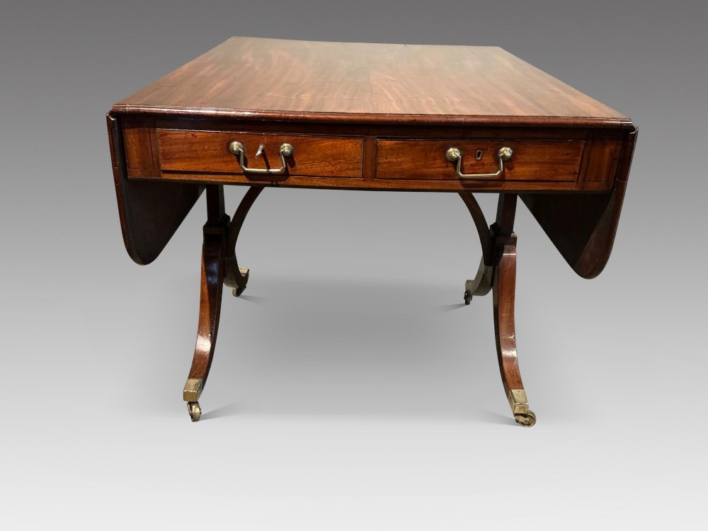 An antique Georgian mahogany sofa table with two drop leaves, two front drawers with brass handles, and splayed legs resting on brass castors. View II
