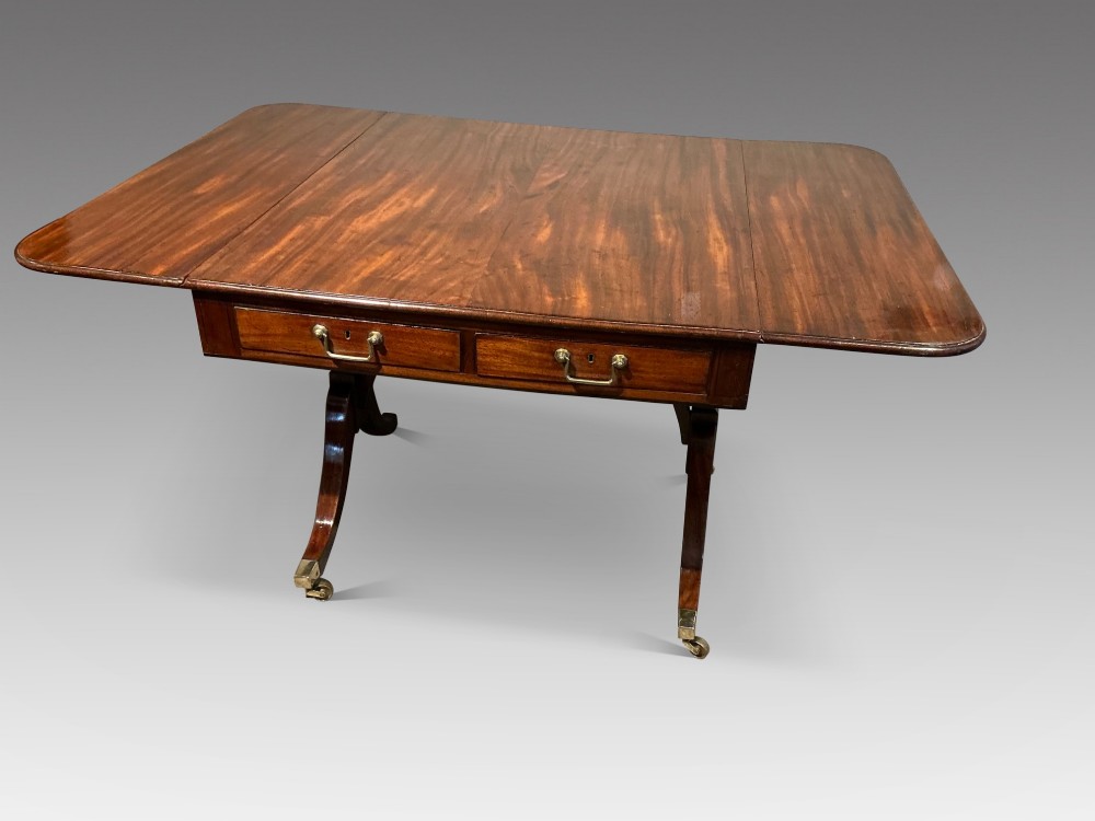 An antique Georgian mahogany sofa table with two drop leaves, two front drawers with brass handles, and splayed legs resting on brass castors. View III