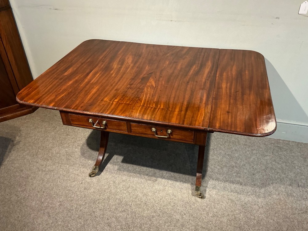 An antique Georgian mahogany sofa table with two drop leaves, two front drawers with brass handles, and splayed legs resting on brass castors. View IV