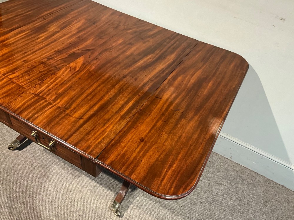 An antique Georgian mahogany sofa table with two drop leaves, two front drawers with brass handles, and splayed legs resting on brass castors. View V