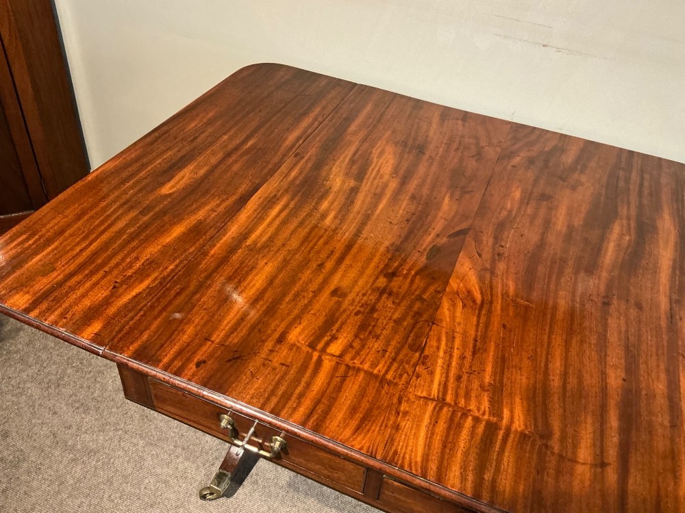 An antique Georgian mahogany sofa table with two drop leaves, two front drawers with brass handles, and splayed legs resting on brass castors. View VI
