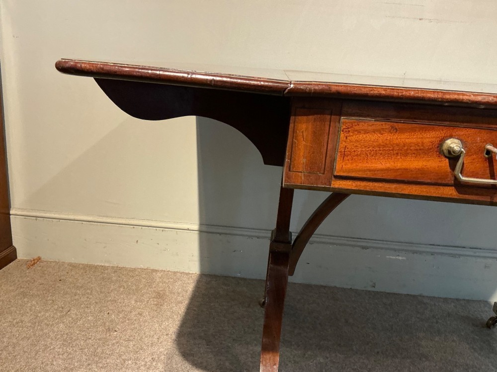 An antique Georgian mahogany sofa table with two drop leaves, two front drawers with brass handles, and splayed legs resting on brass castors. View VII