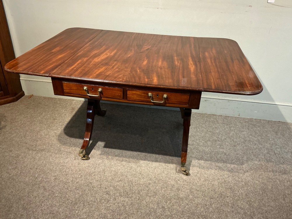 An antique Georgian mahogany sofa table with two drop leaves, two front drawers with brass handles, and splayed legs resting on brass castors. View IX
