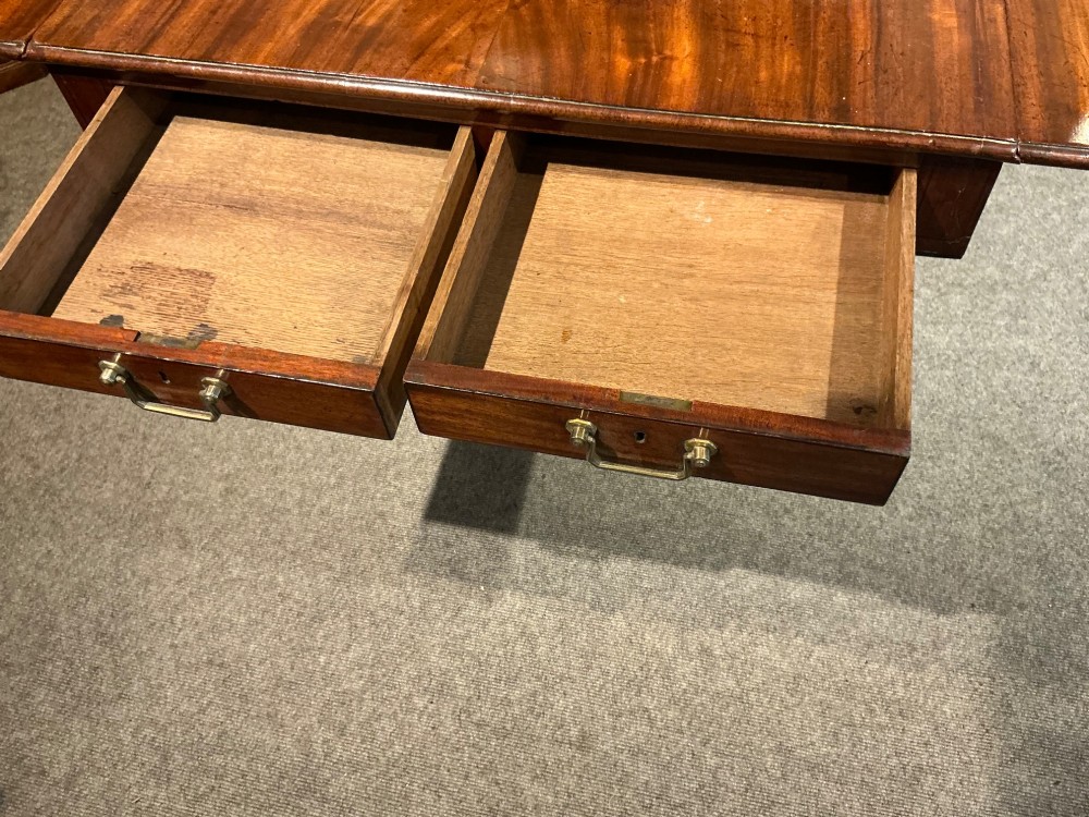 An antique Georgian mahogany sofa table with two drop leaves, two front drawers with brass handles, and splayed legs resting on brass castors. View X