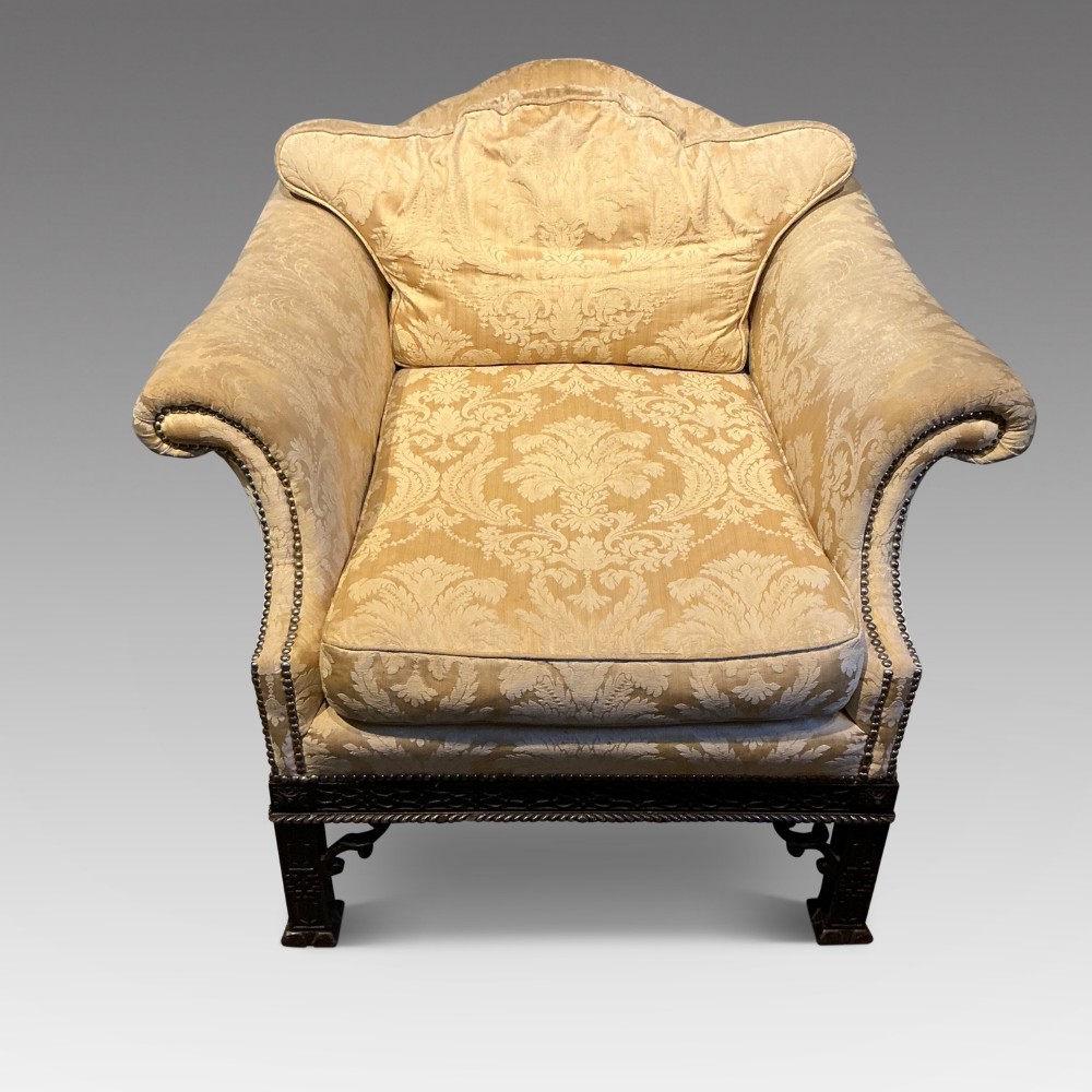 A deep upholstered armchair with gold damask fabric, broad scroll arms with brass stud trim, and carved dark timber legs.