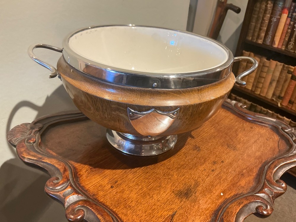 An Edwardian turned oak bowl featuring a silver-plated rim, twin loop handles, a pedestal foot, a blank shield cartouche, and a white ceramic liner. View VI