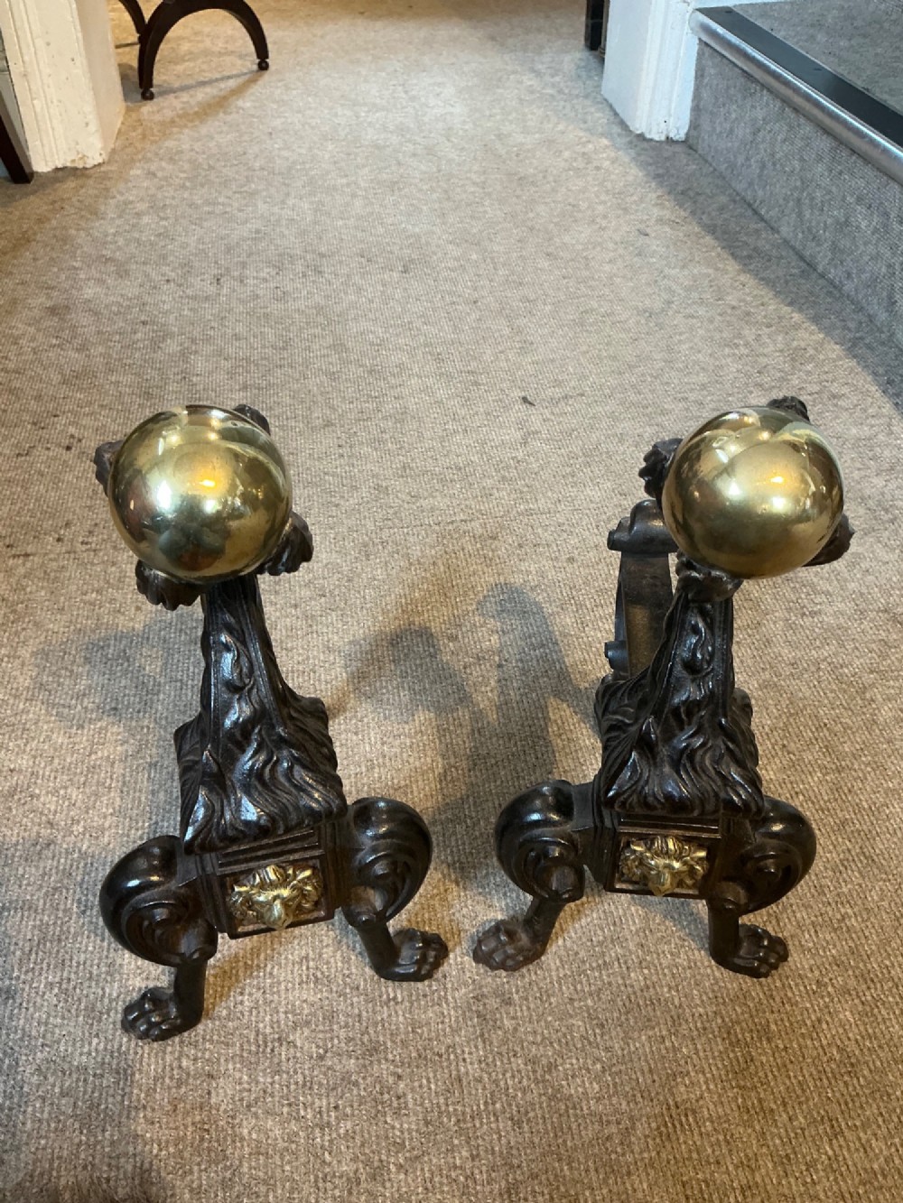 A pair of antique cast iron and brass andirons featuring spherical brass finials, lion mask central motifs, scrolled legs, and claw feet.
