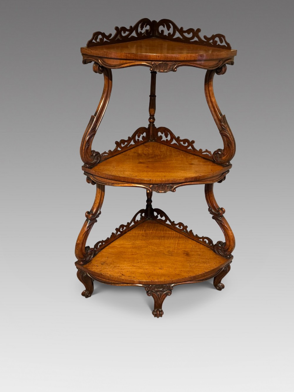 A three-tier antique corner whatnot in warm brown polished wood, featuring pierced fretwork galleries, serpentine supports, and carved feet.