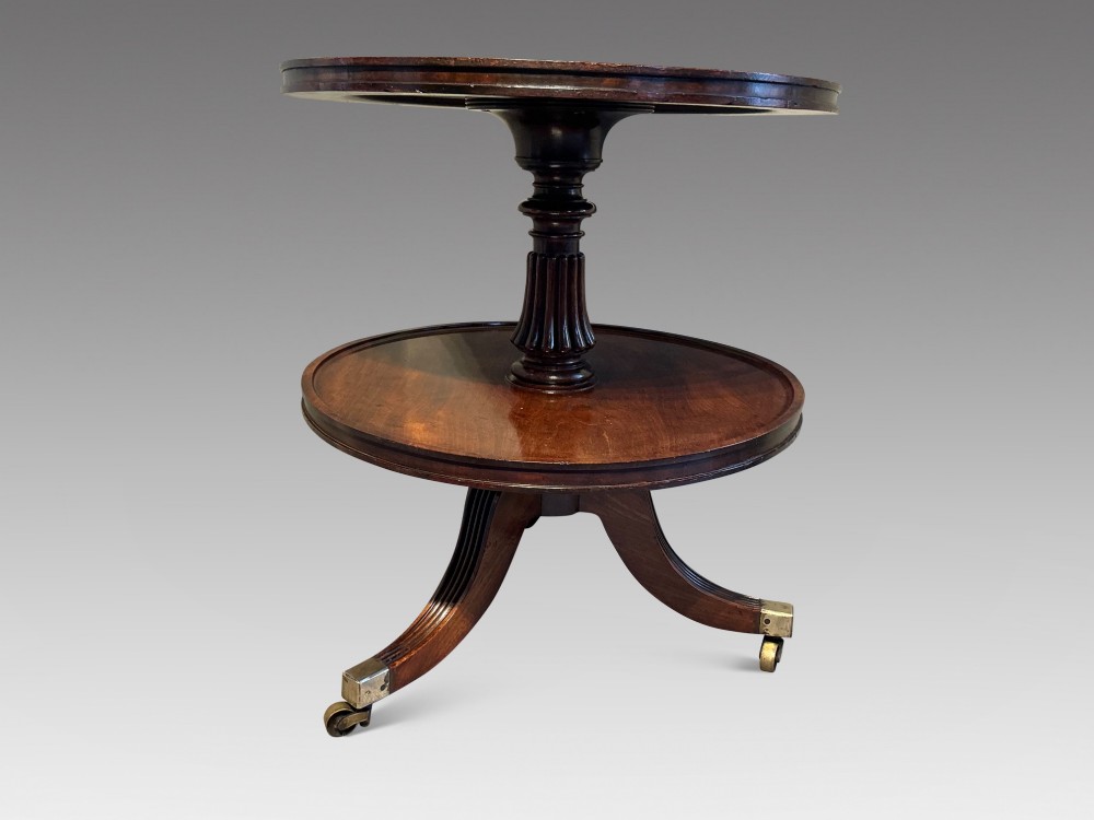 A two-tier dark mahogany Regency dumb waiter featuring a turned central column, reeded tripod legs, and brass castors.