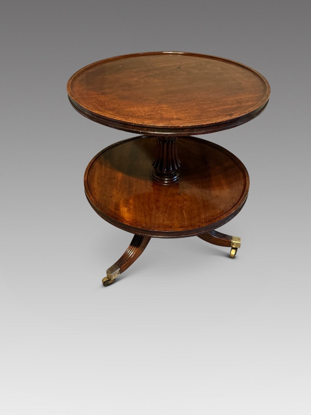 A two-tier dark mahogany Regency dumb waiter featuring a turned central column, reeded tripod legs, and brass castors. View II