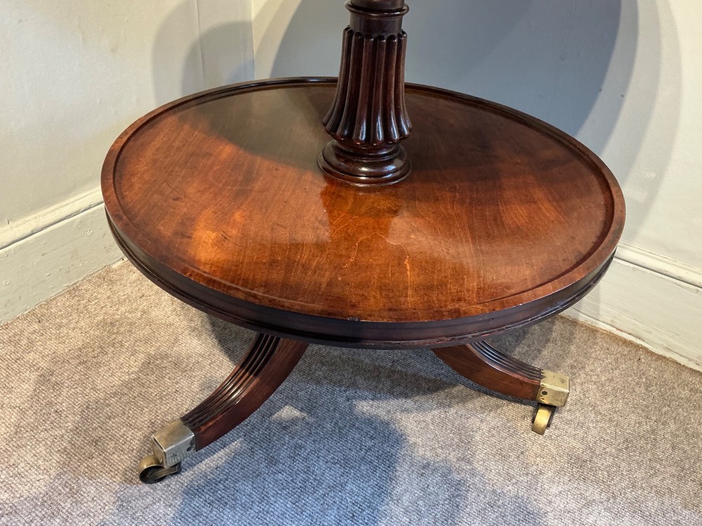 A two-tier dark mahogany Regency dumb waiter featuring a turned central column, reeded tripod legs, and brass castors. View V