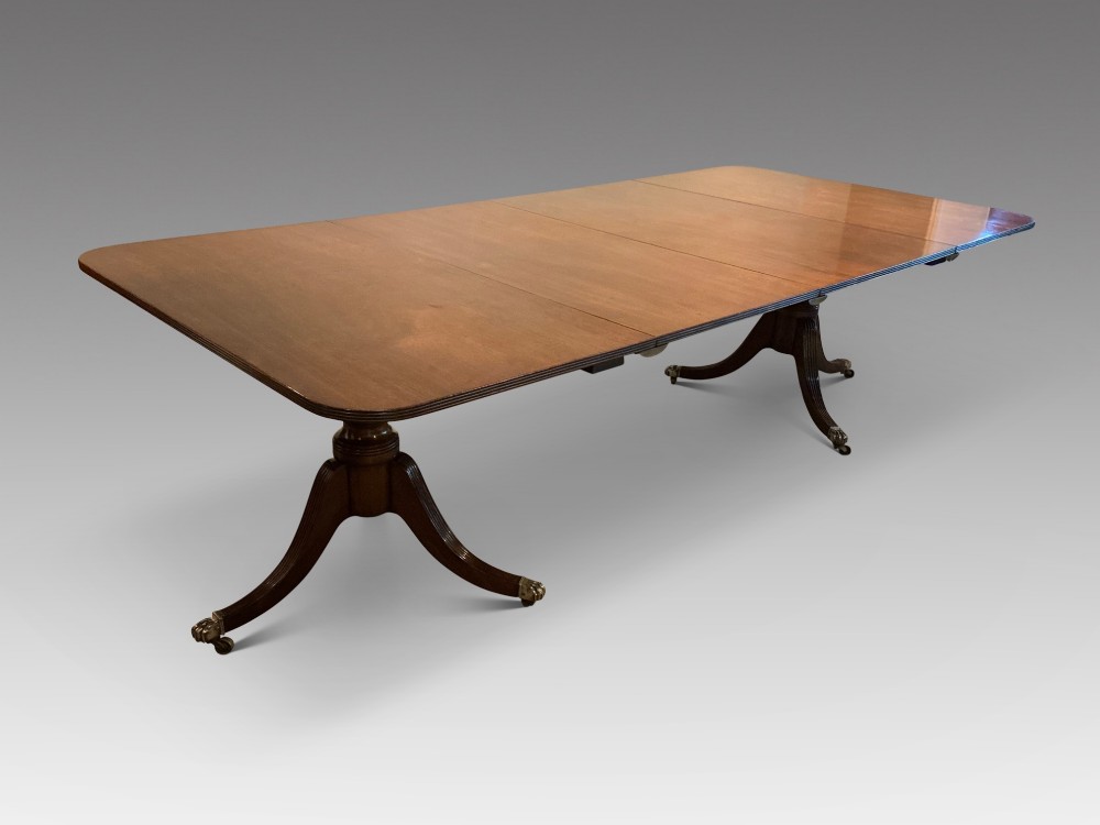 An antique mahogany twin pedestal dining table with rounded corners, extending leaves, turned supports, and splayed legs with brass castors.