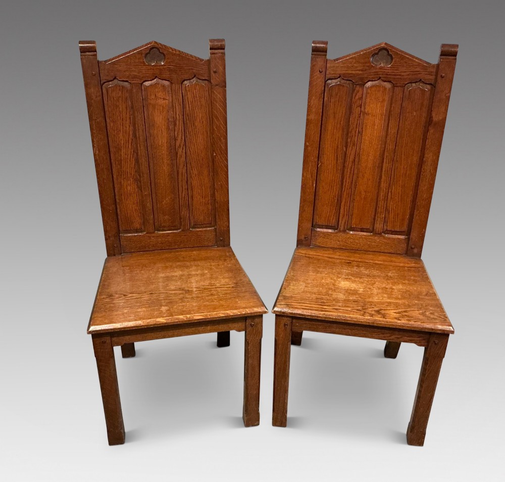 A pair of solid oak hall chairs featuring carved Gothic arches, trefoil back detailing, and flat wood seats.
