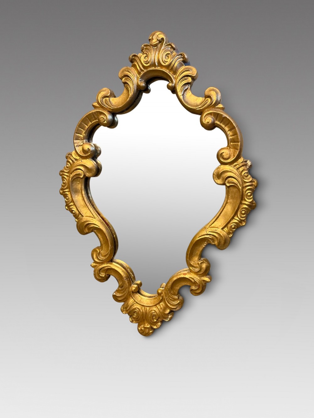 A cartouche-shaped wall mirror featuring a carved gold gilt frame with ornate C-scrolls and decorative flourishes.