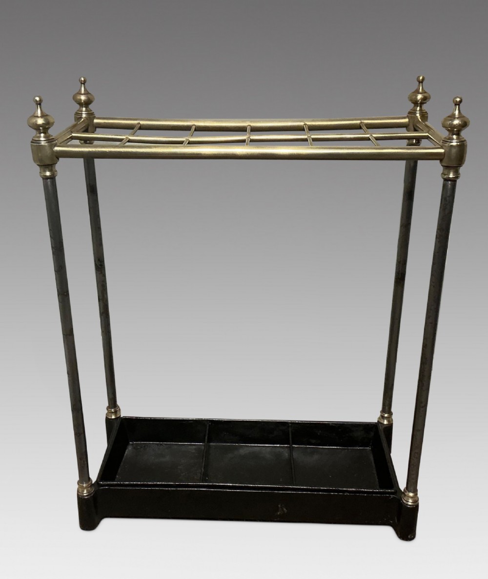 An antique brass and steel stick stand featuring a six-section top grid, urn finials, tubular uprights, and a black cast iron drip tray base.