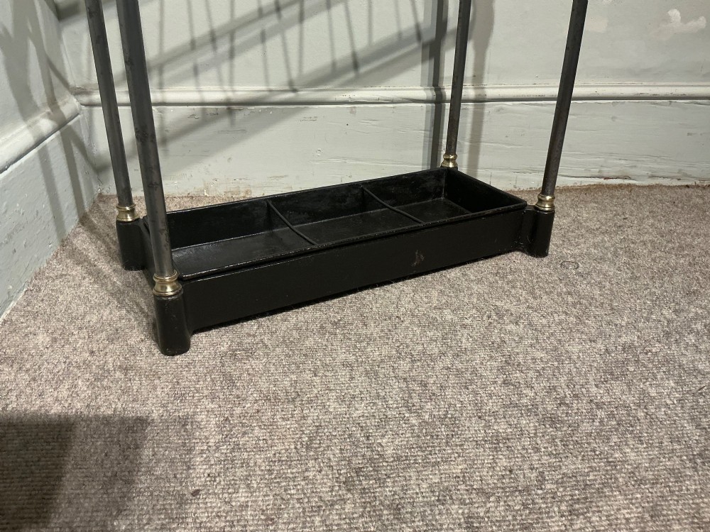 An antique brass and steel stick stand featuring a six-section top grid, urn finials, tubular uprights, and a black cast iron drip tray base. View III