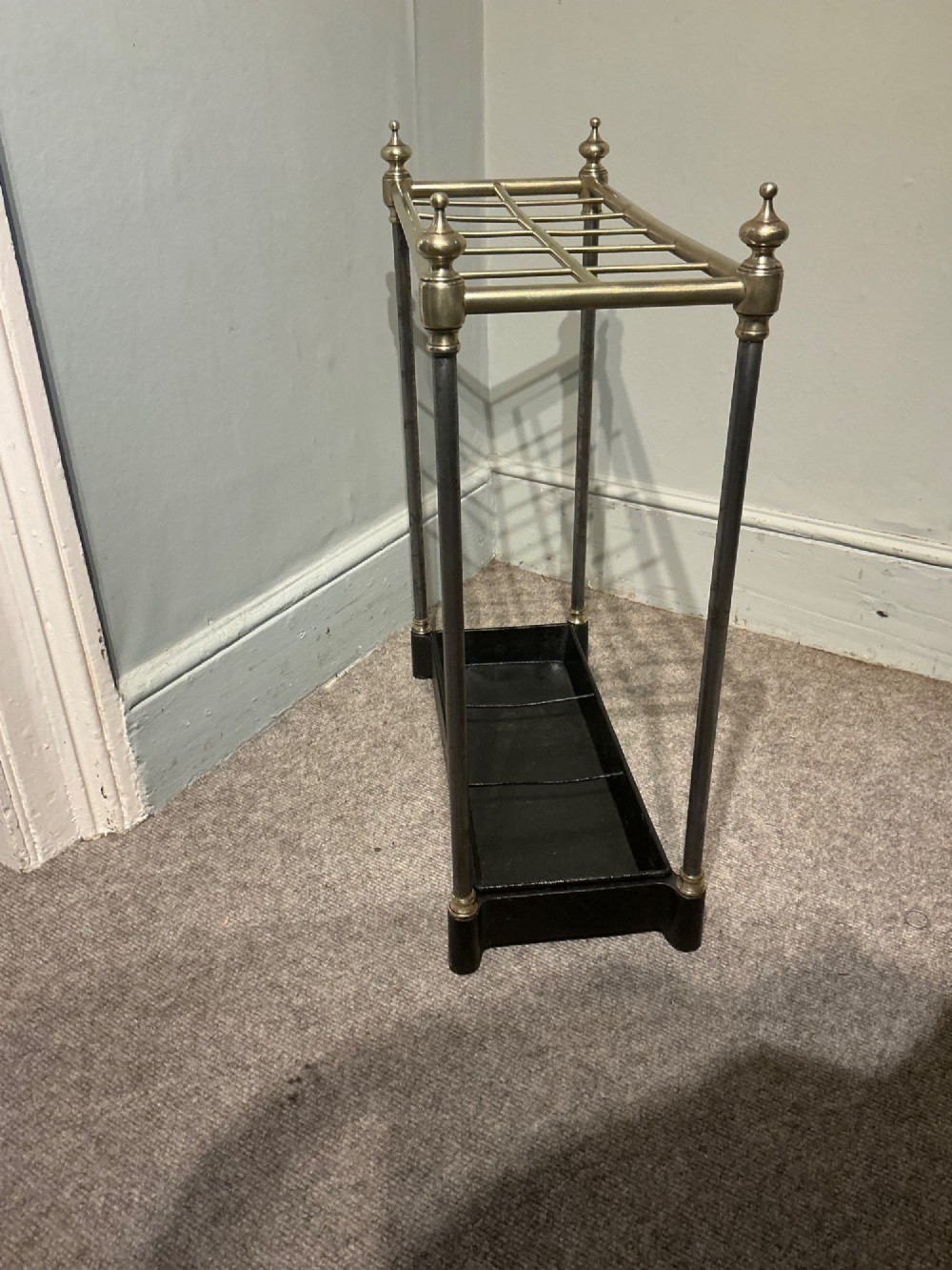 An antique brass and steel stick stand featuring a six-section top grid, urn finials, tubular uprights, and a black cast iron drip tray base. View V